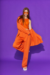 Young fashion woman in orange pants orange top orange shirt on violet background. Platform slides sandals, orange sunglasses.