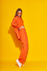 Young fashion woman in orange pants orange top orange shirt on yellow background. Platform slides sandals, orange sunglasses. - 632124640