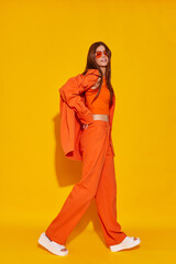 Young fashion woman in orange pants orange top orange shirt on yellow background. Platform slides sandals, orange sunglasses.