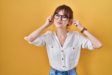 Young beautiful woman wearing casual shirt over yellow background smiling pulling ears with...