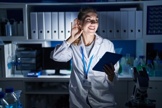 Beautiful blonde woman working at scientist laboratory late at night smiling with hand over ear listening an hearing to rumor or gossip. deafness concept.
