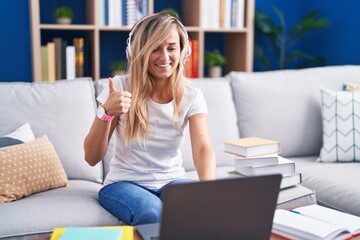 Obraz premium Young blonde woman studying using computer laptop at home doing happy thumbs up gesture with hand. approving expression looking at the camera showing success.