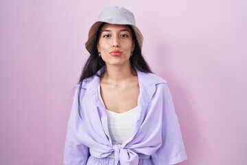 Young hispanic woman standing over pink background wearing hat looking at the camera blowing a kiss...