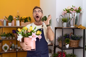 Middle age man with beard working at florist shop holding plant celebrating victory with happy smile and winner expression with raised hands