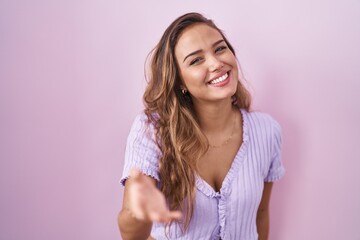 Young hispanic woman standing over pink background smiling cheerful offering palm hand giving assistance and acceptance.