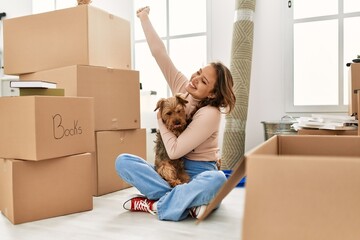Young beautiful hispanic woman hugging dog sitting on floor with cheerful expression at new home