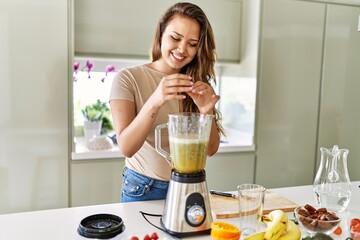 Young beautiful hispanic woman preparing vegetable smoothie with blender at the kitchen