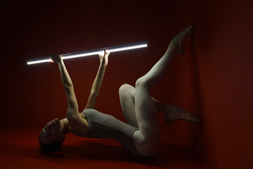 ballerina in bodysuit and tights poses stretching in a dark photo studio on a red background with a...