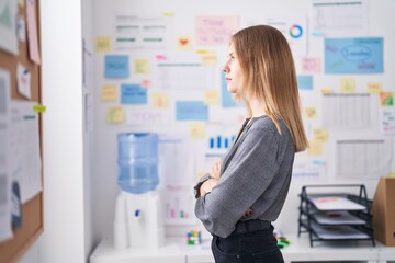 Young blonde woman business worker looking throw the window with serious face and arms crossed gesture at office