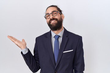Hispanic man with beard wearing suit and tie smiling cheerful presenting and pointing with palm of hand looking at the camera.