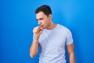 Young hispanic man standing over blue background feeling unwell and coughing as symptom for cold or...
