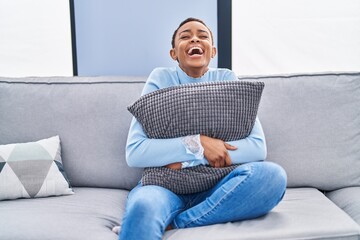 African american woman sitting on the sofa at home hugging pillow smiling and laughing hard out loud because funny crazy joke.