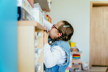 Inquisitive girl with special needs wearing eyeglasses taking box with game from shelves at...