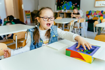 Excited girl with down syndrome sitting at desk smiling and playing at class. Developmental...