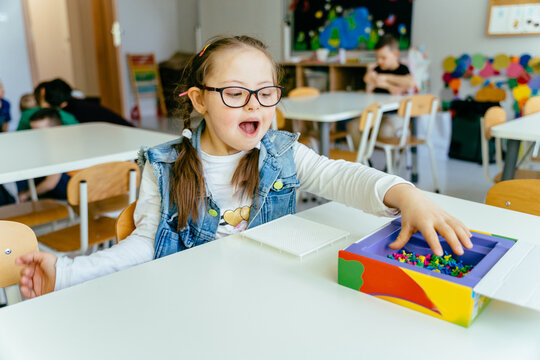 Excited girl with down syndrome sitting at desk smiling and playing at class. Developmental activities with children. People with disability.
