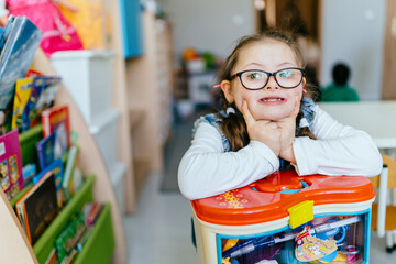 Cute adorable schoolgirl with down syndrome sitting in he classroom showing big smile. Back to school. Happy little girl ready for primary school. - Powered by Adobe