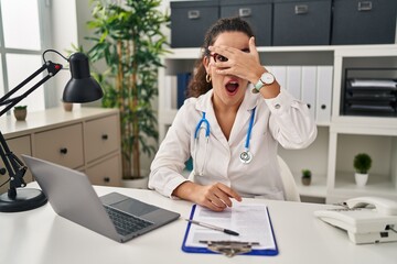 Young hispanic woman wearing doctor uniform and stethoscope peeking in shock covering face and eyes with hand, looking through fingers with embarrassed expression.