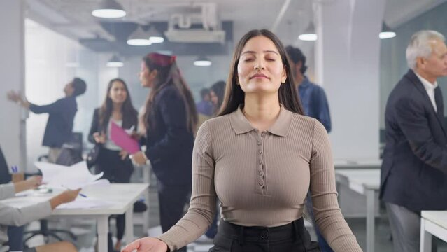 A young Asian Indian corporate woman employee sitting quietly with closed eyes in a meditative pose middle of a busy chaotic and messy office where people or coworkers throwing papers, fighting