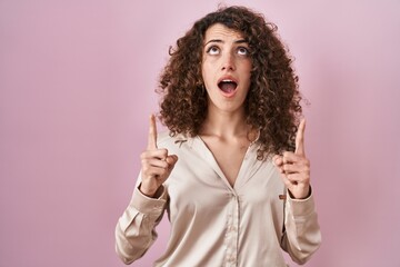 Hispanic woman with curly hair standing over pink background amazed and surprised looking up and pointing with fingers and raised arms.