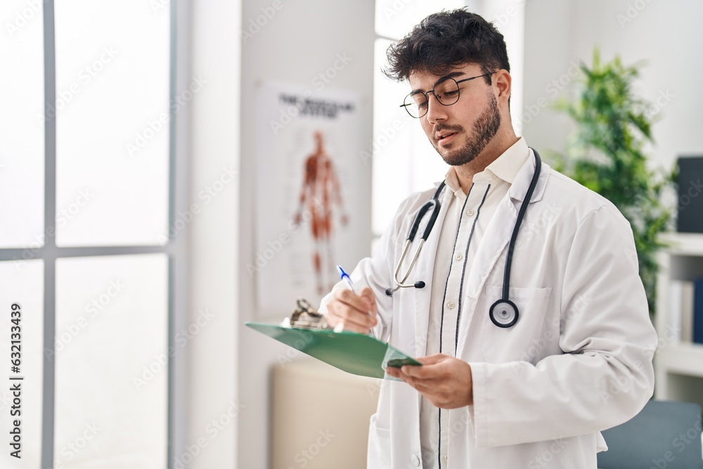 Sticker young hispanic man wearing doctor uniform writing medical report at clinic