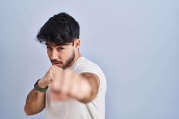 Hispanic man with beard standing over white background punching fist to fight, aggressive and angry...
