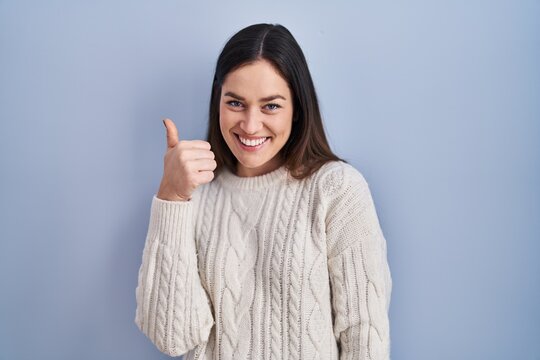 Young brunette woman standing over blue background doing happy thumbs up gesture with hand. approving expression looking at the camera showing success.