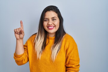 Young hispanic woman standing over isolated background showing and pointing up with finger number one while smiling confident and happy.