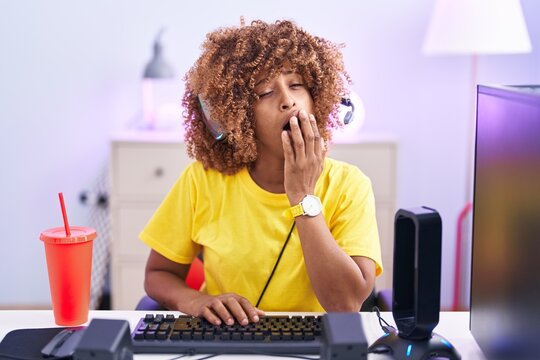 Young hispanic woman with curly hair playing video games wearing headphones bored yawning tired covering mouth with hand. restless and sleepiness.