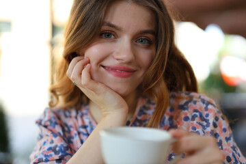 A cheerful young woman enjoying a hot coffee in a summer morning. She is holding a white cup and smiling while sitting at an outdoor cafe with green plants.