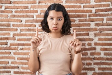 Young hispanic woman standing over bricks wall pointing up looking sad and upset, indicating...