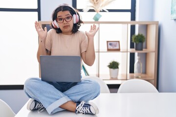 Young hispanic woman using laptop sitting on the table wearing headphones clueless and confused expression with arms and hands raised. doubt concept.
