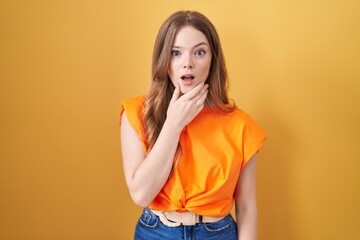 Caucasian woman standing over yellow background looking fascinated with disbelief, surprise and amazed expression with hands on chin
