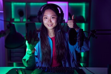 Young asian woman playing video games with smartphone doing happy thumbs up gesture with hand. approving expression looking at the camera showing success.