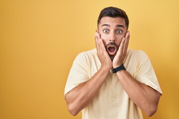 Handsome hispanic man standing over yellow background afraid and shocked, surprise and amazed expression with hands on face