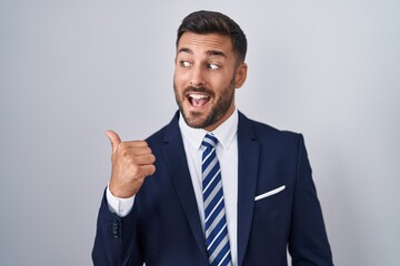 Handsome hispanic man wearing suit and tie smiling with happy face looking and pointing to the side with thumb up.