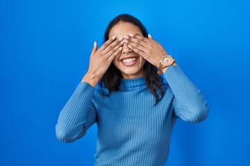 Young brazilian woman standing over blue isolated background covering eyes with hands smiling...