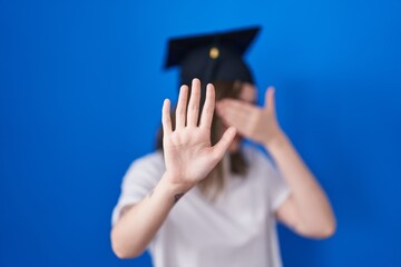 Blonde caucasian woman wearing graduation cap covering eyes with hands and doing stop gesture with...