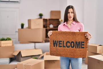 Young hispanic woman holding welcome doormat at new home in shock face, looking skeptical and...