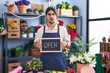 Hispanic man with long hair working at florist holding open sign in shock face, looking skeptical and sarcastic, surprised with open mouth