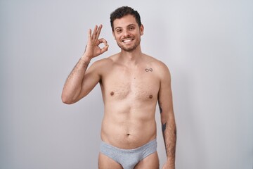 Fototapeta premium Young hispanic man standing shirtless wearing underware smiling positive doing ok sign with hand and fingers. successful expression.