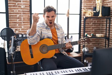 Young hispanic man playing classic guitar at music studio annoyed and frustrated shouting with...