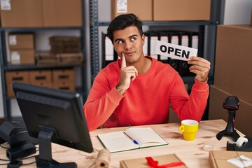 Hispanic man working at small business ecommerce holding open banner serious face thinking about...