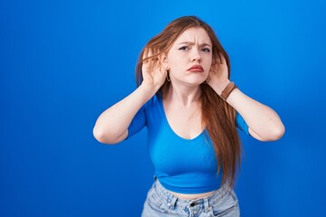 Obraz na płótnie Canvas Redhead woman standing over blue background trying to hear both hands on ear gesture, curious for gossip. hearing problem, deaf