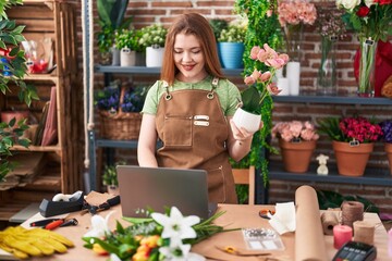 Young redhead woman florist using laptop holding plant at flower shop