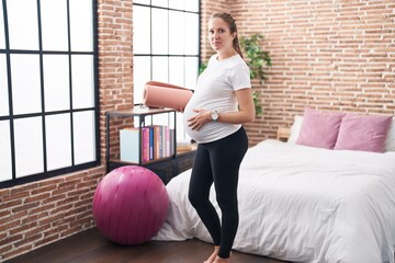 Young pregnant woman holding pilate mat at bedroom relaxed with serious expression on face. simple...