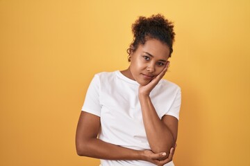 Obraz premium Young hispanic woman with curly hair standing over yellow background thinking looking tired and bored with depression problems with crossed arms.