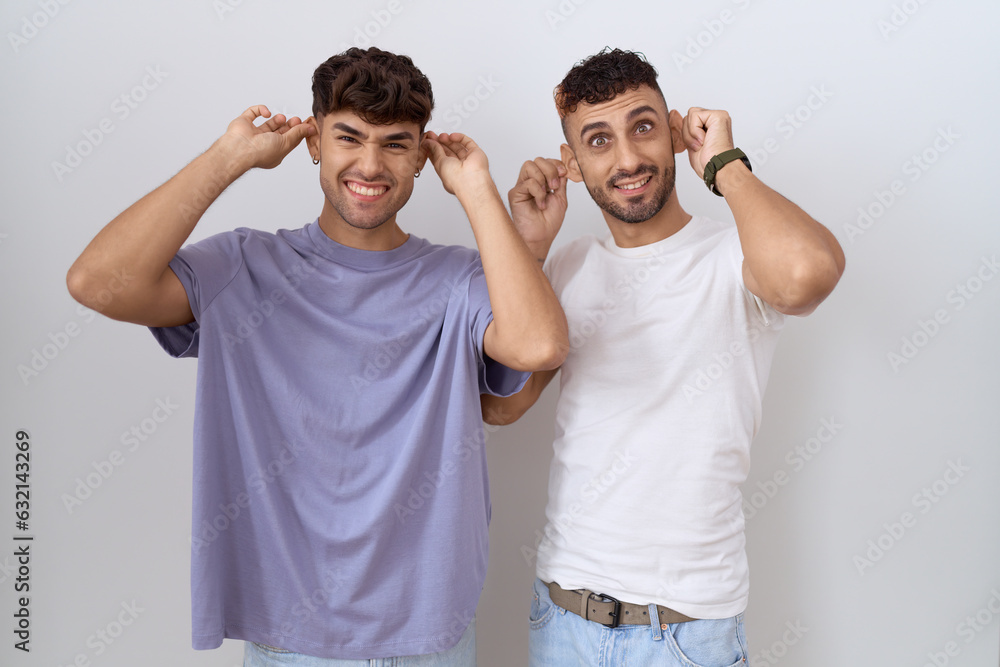 Sticker Homosexual gay couple standing over white background smiling pulling ears with fingers, funny gesture. audition problem