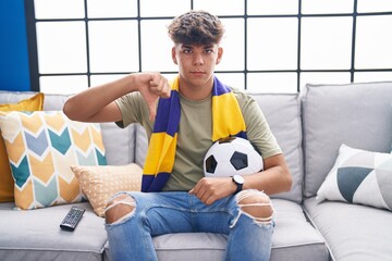 Hispanic teenager sitting on the sofa watching football match with angry face, negative sign...