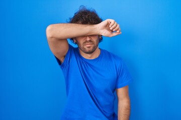 Hispanic young man standing over blue background covering eyes with arm, looking serious and sad....