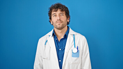 Young hispanic man doctor standing with serious expression over isolated blue background